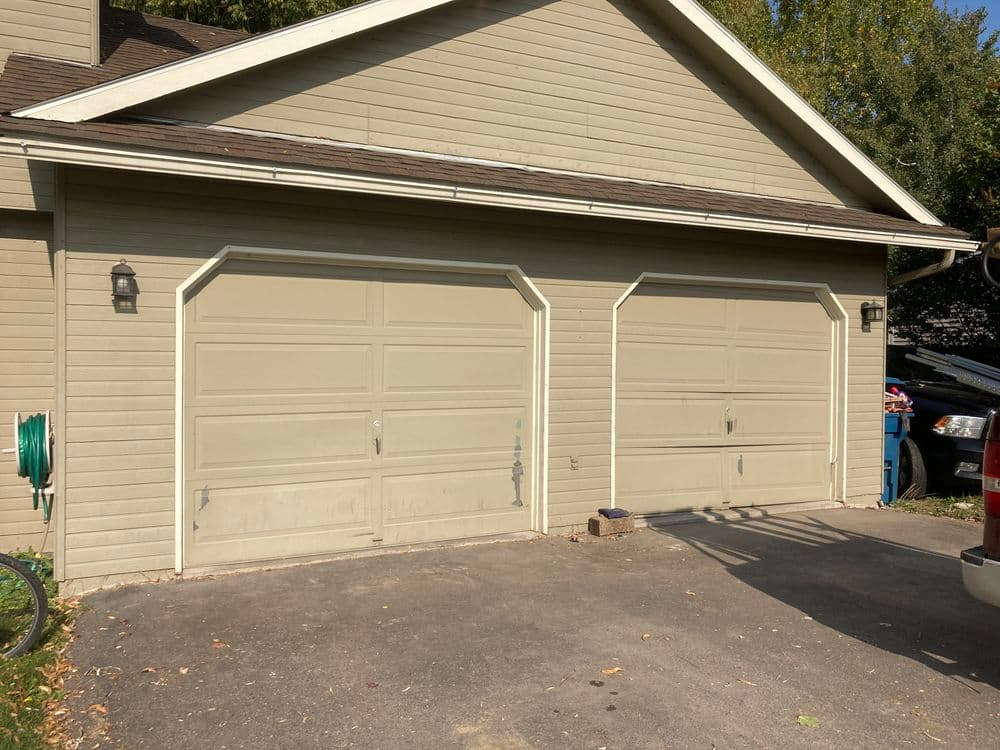Two beige garage doors on a suburban house with a paved driveway and greenery.