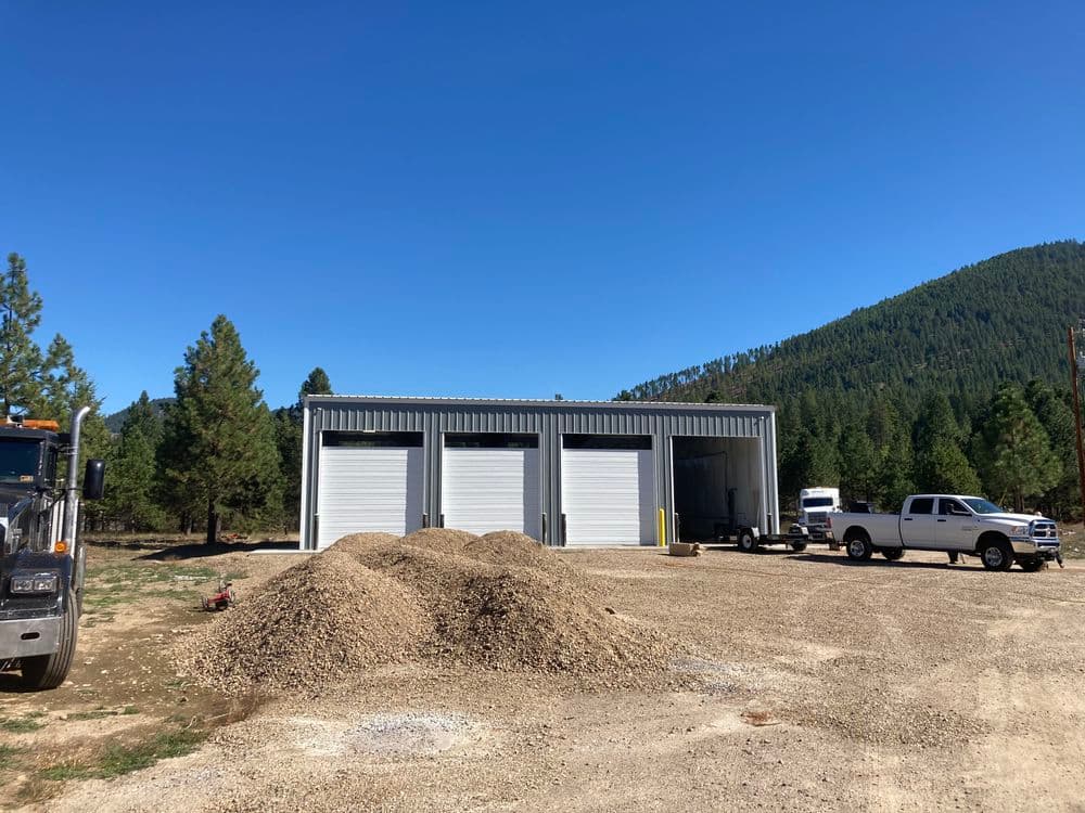 Steel storage building with three roll-up doors, surrounded by gravel and pine trees.