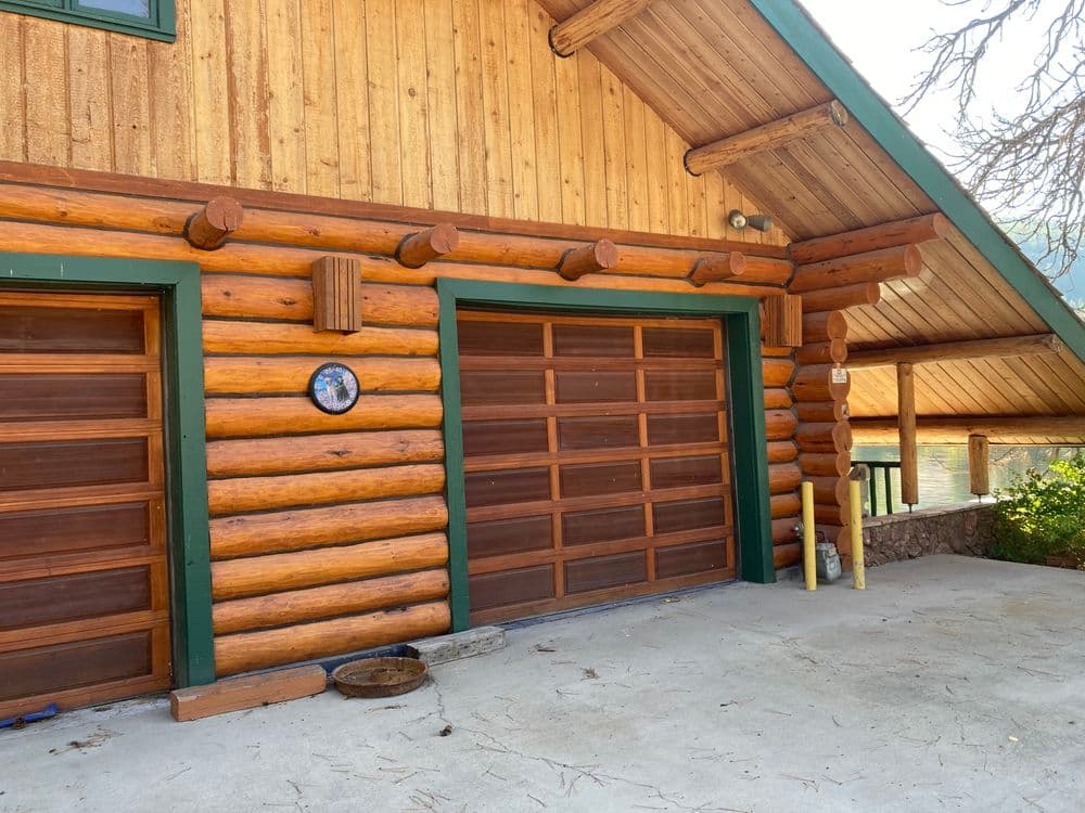 Log cabin garage with wooden doors and rustic exterior, surrounded by nature.