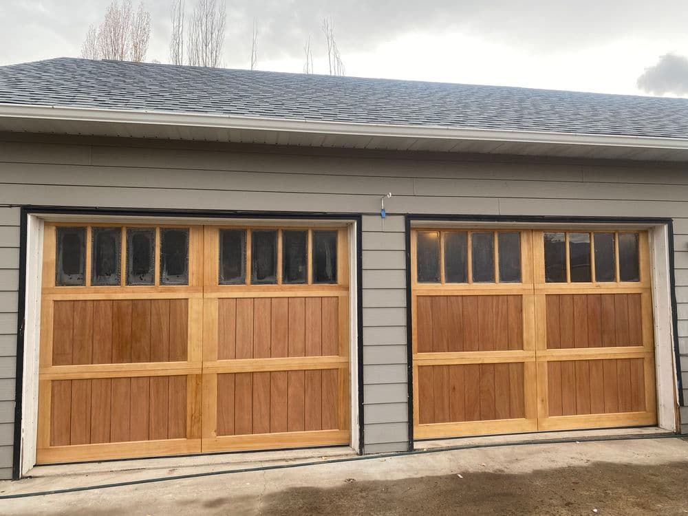 Wooden garage doors with glass panels on a gray house exterior. Modern design and craftsmanship.