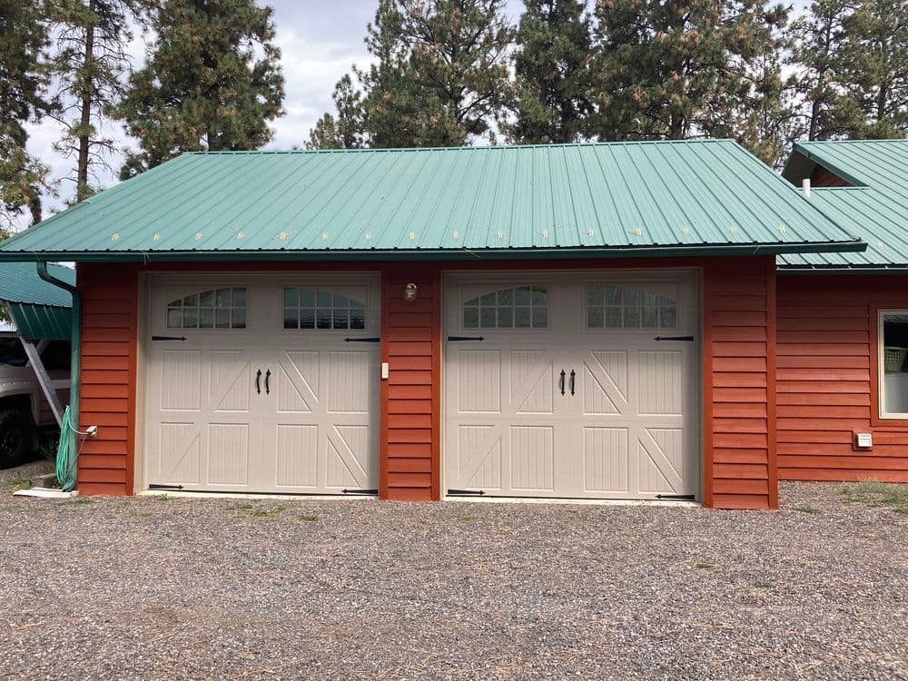 Red wooden garage with beige double doors and a green metal roof, set in a wooded area.