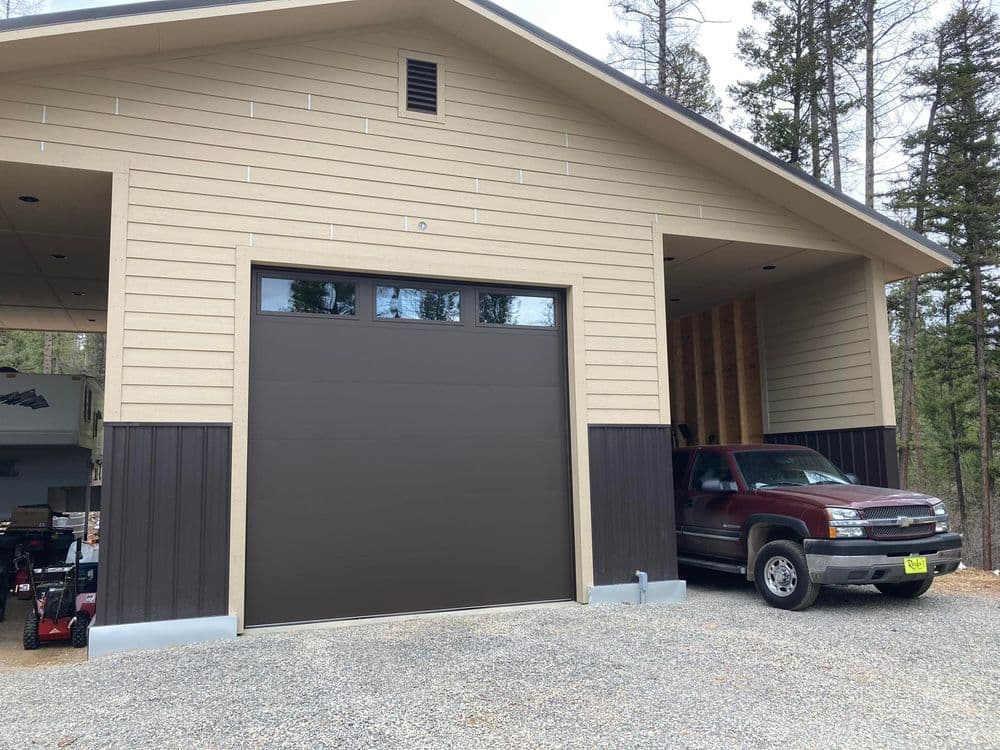 Modern garage with brown door and red truck parked outside, surrounded by trees.