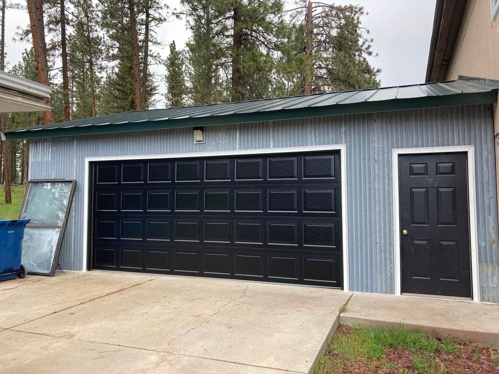 Metal garage with black doors and a side entry, surrounded by trees and concrete driveway.
